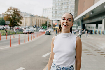 Happy caucasian girl looking into camera while posing in city centre. Refined short-haired woman in white shirt and jeans enjoying sunny summer morning