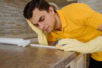 Young man in a yellow t-shirt and yellow gloves is tired after cleaning the wash basin sink in the...