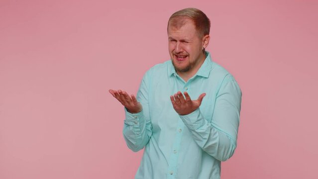 Upset disappointed handsome stylish man in blue shirt wipes tears and cries from despair, being sad because of unfair things. Young adult guy. Indoors studio shot isolated alone on pink background