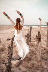 Model in boho style in a white long dress and silver jewelry on the beach. Her hair is braided, and there are many bracelets on her arms. © svetograph