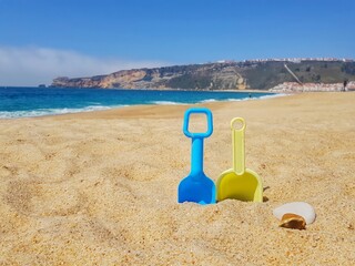 Fototapeta na wymiar Sandy beach with coastal mountain and blue sky background. Two blue and yellow toy shovels stuck in the sand.