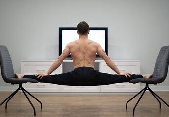 Flexible man keep balance in split between two chairs watching TV. Concept of willpower, motivation and passion