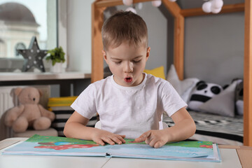 Cute little boy reading book at table in room