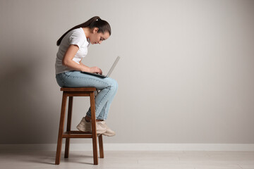 Young woman with poor posture using laptop while sitting on stool near grey wall, space for text - Powered by Adobe
