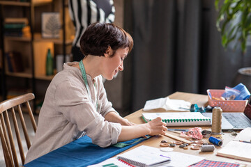 Fototapeta premium Very pretty female fashion designer while sew some fabrics at her workplace in the tailoring atelier looking up to the camera and smiling large