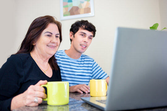 Happy mother and son sitting at kitchen table at home and using laptop to browse the internet or make online shopping order.