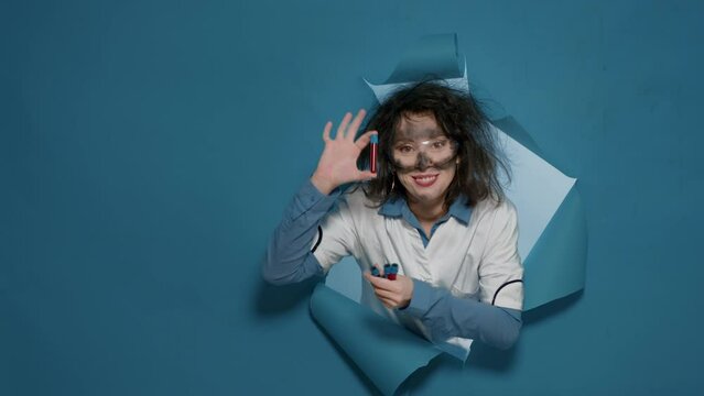 Crazy silly scientist grinning and showing test tubes on camera, doing foolish mad facial expressions. Goofy sunny woman acting insane and amusing, having messy insane hair in studio.