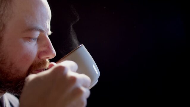 SLOW MOTION BACKLIT - A man drinks from a steaming cup of hot beverage