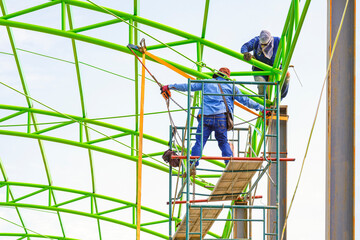 Asian foreman and construction worker on scaffolding are repairing metal roof structure of industrial building in construction site