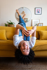 Happy African American woman lying upside down on the couch texting using mobile phone. Checking social media. Vertical. © Daniel