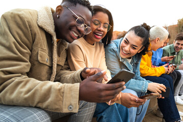 Happy multiracial teen college students having fun using mobile phone together in campus outdoors.