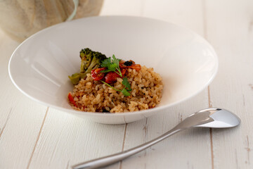 Risotto in a white bowl on a white wooden country feeling table, topped with herbs, broccoli and oiled tomatoes. 