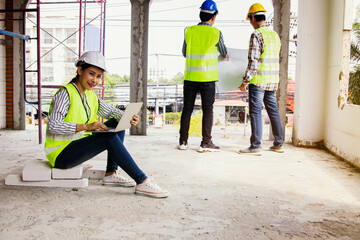 Architect businessman beautiful asian woman wearing helmet and yellow vest sits with laptop examining construction site reports two male workers standing behind building's construction site consulting