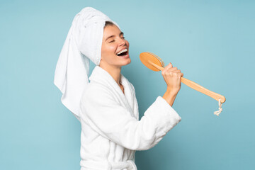 Young funny woman singing after shower in bathrobe and towel over head isolated on blue - obrazy, fototapety, plakaty