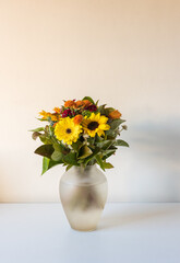 Vertical close up of yellow and orange flowers in glass vase including gerbera daisies and sunflowers against beige wall (selective focus)