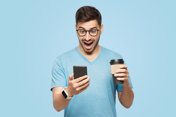 Handsome man with cup of coffee, looking at phone screen with shocked face