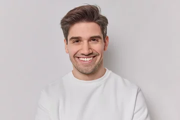  Portrait handsome man with dark hairstyle bristle and toothy smile dressed in white sweatshirt feels very glad poses indoor. Pleased European guy being in good mood smiles positively. Emotions concept © Wayhome Studio