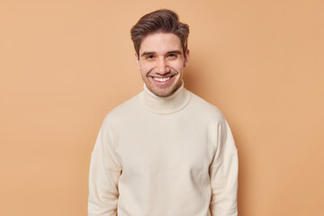 Portrait of handsome young man with dark hair toothy smile looks gladfully at camera wears casual white turtleneck isolated over brown background. Smiling adult male model being in good mood