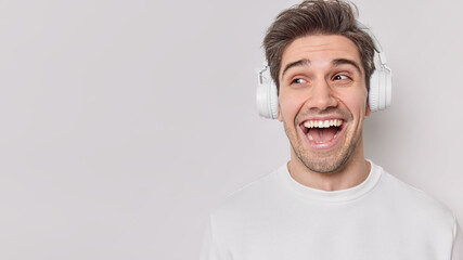 Horizontal studio shot of happy brunet adult man wears wireless headphones on ears listens favorite music during free time poses against white background blank space for your promotional content