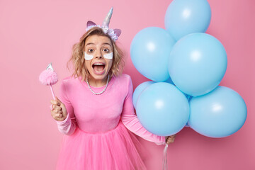 Obraz na płótnie Canvas Happy amazed young European woman exclaims loudly applies beauty patches under eyes wears dress holds bunch of inflated bluue balloons isolated over pink background. Festive occasion concept