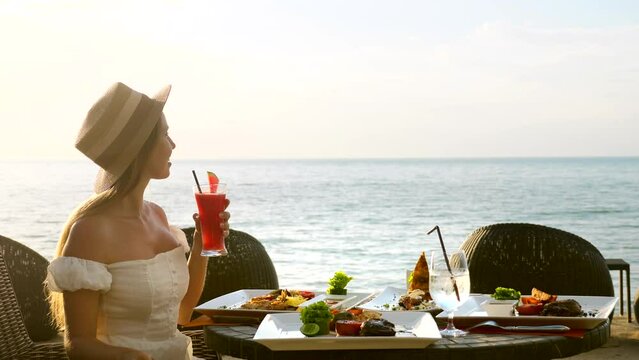 Young woman have romantic breakfast in sunrise at resort fine dining restaurant outdoor enjoy vacation looking at sea. Beautiful girl wear hat hold fruit cocktail. Healthy food drink for breakfast.
