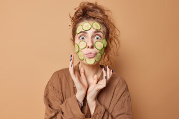Skin care and wellness concept. Surprised woman has messy hair applies cucumber slices on face to...