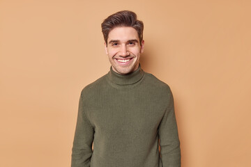 Portrait of handsome young man with dark hair smiles happily expresses positive emotions dressed in casual turtleneck hears good nes isolated over brown background glad to meet old best friend