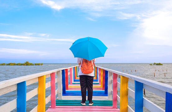 Rear view of female tourist with backpack holding umbrella on the old multicolored wooden bridge at sea viewpoint against clouds on blue sky background