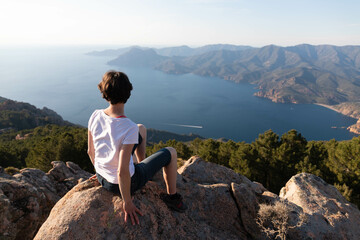 Fototapeta na wymiar Young woman sitting alone in front of a breathtaking panoramic landscape at sunset in Corsica, meditation position