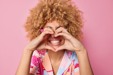 Positive curly haired young woman makes heart gesture over toothy smile has good romantic mood...