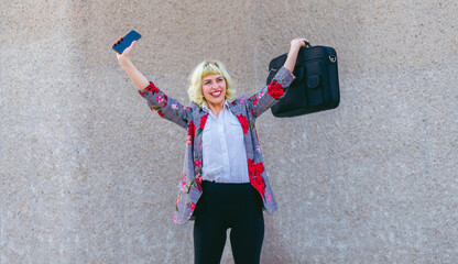 El éxito de la mujer en el mundo laboral,  celebración de los objetivos y metas , mujer feliz con los brazos levantados sujetando un maletín y un teléfono móvil  - obrazy, fototapety, plakaty