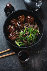 Asian style teriyaki meatballs with soba and scallions served in a black bowl, vertical shot on a dark-brown stone background