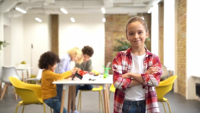 Joyful girl smiling at camera, standing with arms crossed while posing for camera during crafting classes. Kids working on diy robot in the background