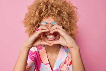 Happy curly haired young woman makes heart gesture over mouth smiles toothily wears transparent spectacles and colorful shirt says I love you isolated over pink background. Body language concept - 502001178