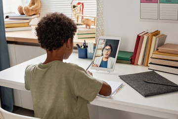 Rear view of black boy sitting at desk in nursery room and making notes while working with tutor