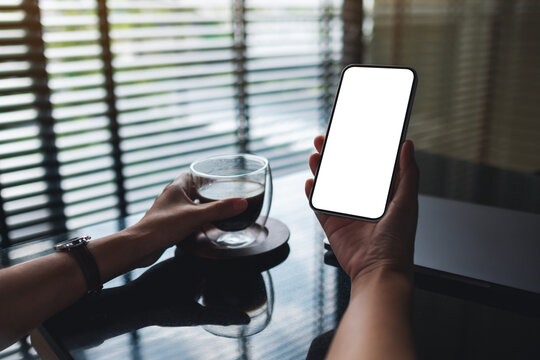 Mockup image of a woman holding mobile phone with blank desktop screen while drinking coffee with laptop on the table