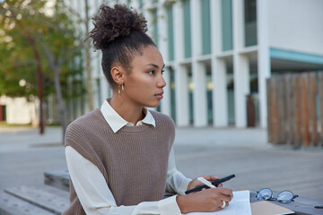 Thoughtful curly haired female student does homework outdoors writes down notes in diary holds mobile phone browses websites wears white shirt and vest focused into distance. Studying concept
