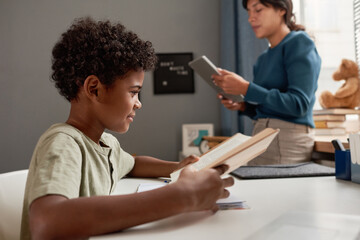 Content curious African American boy sitting at table and reading textbook while teacher using tablet in background