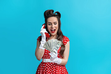 Excited young pinup woman in red polka dot dress holding money, speaking on landline phone on blue...