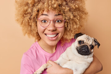 Portrait of happy woman with blonde curly hair embraces pug dog with love carries to veterinary clinic glad to find out her pet is healthy poses against brown studio background. Domestic animals