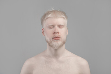 Portrait of young albino man with pale skin, white hair and beard posing with closed eyes against grey background