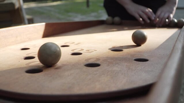 Woman is trying to roll wooden balls down tilted board into the holes with prize points. Hands pushing chip in wooden board game, balls do not fall into pockets, close-up.
