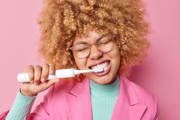 Horizontal shot of curly young woman brushes teeth every morning tries to be always healthy uses electric toothbrush wears transparent spectacles formal clothes isolated on pink wall. Tooth brushing