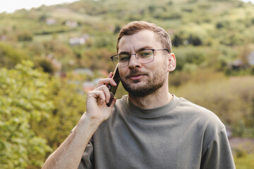 Handsome confident young adult man in eyeglasses stands outdoors in the countryside and talks on the mobile phone.