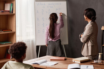 Brown-haired girl writing down on whiteboard while solving math problem under control of teacher at lesson