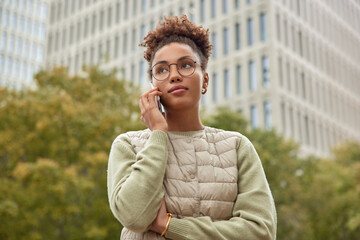 Pensive curly haired woman has telephone conversation focused into distance with thoughtful...