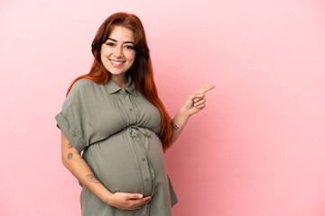 Young redhead caucasian woman isolated on pink background pregnant and pointing to the lateral