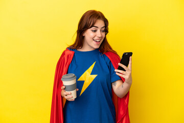 Super Hero redhead woman isolated on yellow background holding coffee to take away and a mobile