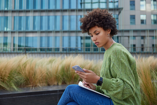 Sideways shot of serious curly haired woman checks schedule online surfs information in internet via mobile phone writes down notes in diary wears green jumper and jeans. Student watches webinar