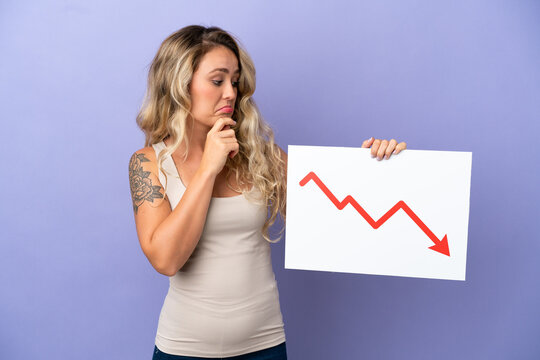 Young Brazilian woman isolated on purple background holding a sign with a decreasing statistics arrow symbol and thinking
